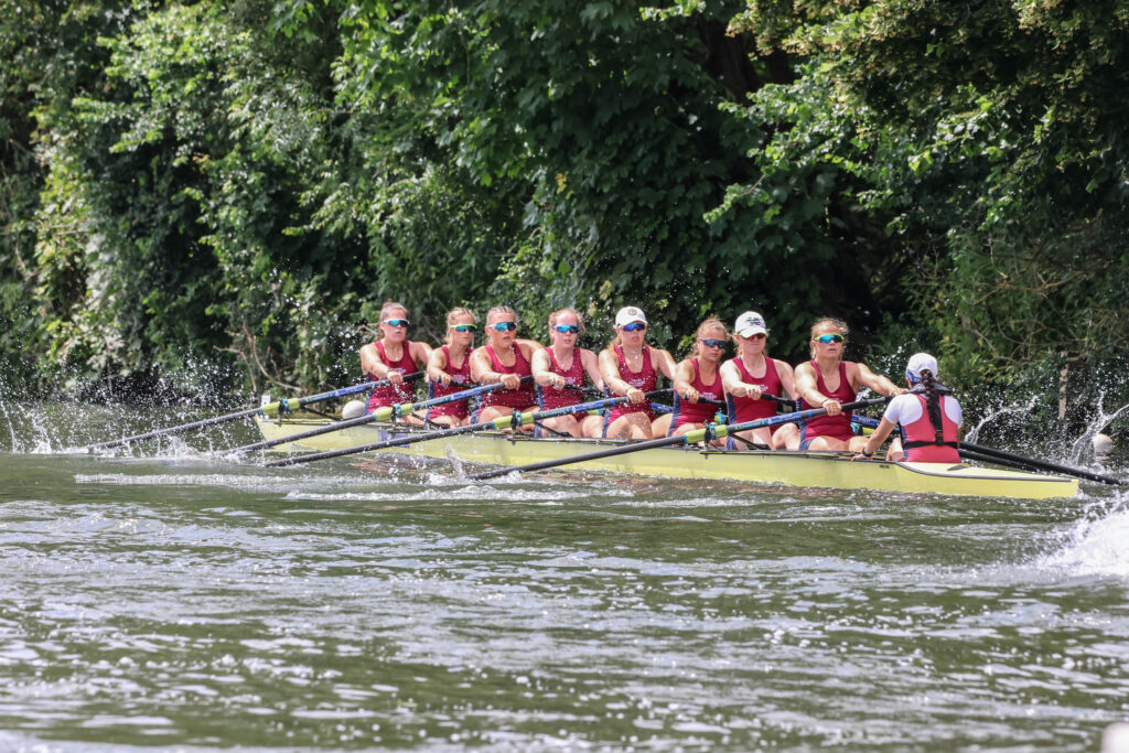 Nine athletes in maroon rowsuits race a yellow eight, rowing away from the camera against a background of trees