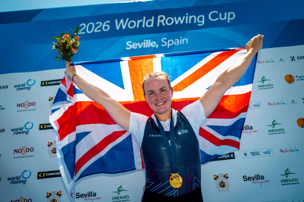 Lauren Henry holds up a Union flag behind her, with a gold medal around her neck and a bunch of flowers. She stands in front of a '2026 World Rowing Cup I' Seville backdrop