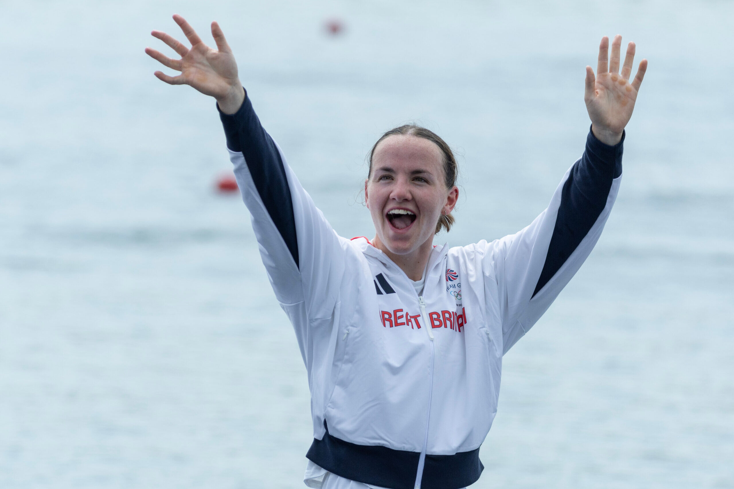Lauren Henry, wearing a white Team GB tracksuit, raises her hands in the air while cheering