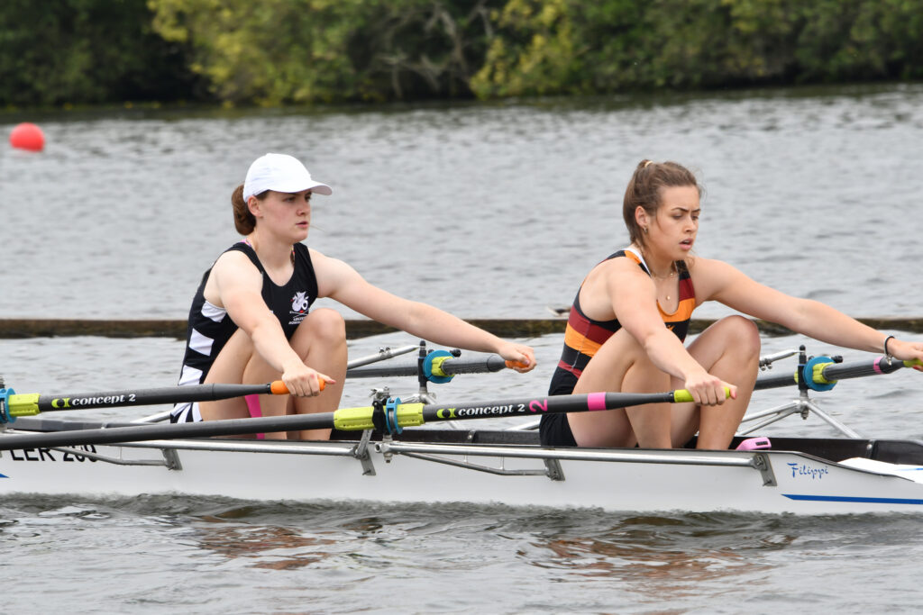 Two young women race a double scull, seen from the side; one wears a black and white rowing suit with a white hat, the other a maroon, orange and black striped suit with no hat